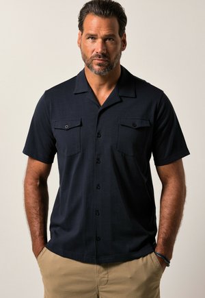 Homme aux cheveux courts et foncés avec une barbe, portant une chemise noire à manches courtes à boutons avec poches poitrine et un pantalon beige sur un fond uni.