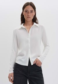 Donna con lunghi capelli castani, che indossa una camicia bianca a maniche lunghe con bottoni e jeans neri, con una mano in tasca, in piedi davanti a uno sfondo neutro.