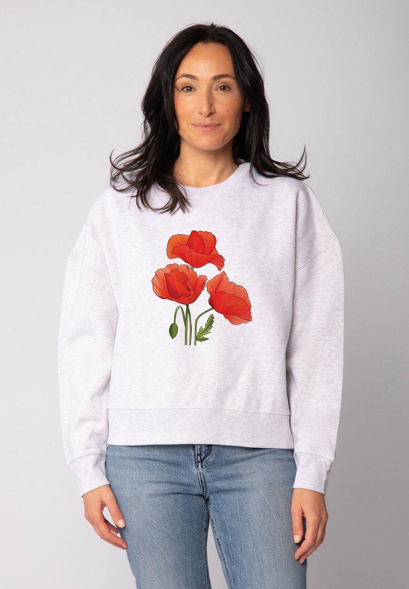 Femme aux cheveux foncés portant un sweat-shirt gris clair avec trois fleurs de coquelicot rouges et un jean bleu, se tenant contre un fond uni.