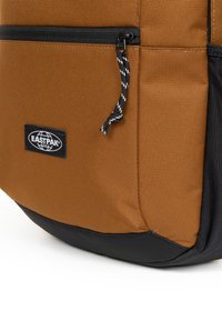 Zaino in tessuto marrone con fondo nero, dotato di una tasca con cerniera e di una toppa con logo del marchio. Presenta una finitura testurizzata e un cursore della cerniera a contrasto.