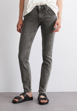 Personne portant un jean skinny gris délavé, une chemise blanche rentrée et des sandales noires à deux brides avec boucles, debout contre un sol gris uni et un mur blanc.