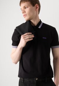 Crna polo majica s ljubičastim i bijelim prugastim ovratnikom; sadrži mali ljubičasti logotip "BOSS" na prsima; lagani materijal.