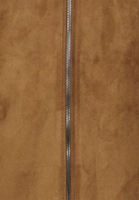 Un material de suede maro, cu un fermoar central argintiu, evidențiind o textură netedă. Designul este simplu și fără prea multe ornamente.