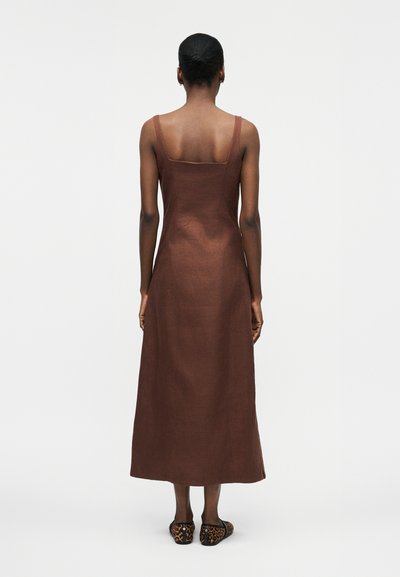 Γυναίκα που στέκεται με την πλάτη στην κάμερα, φορώντας ένα αμάνικο καφέ μίντι φόρεμα και μοκασίνια με λεοπάρ μοτίβο, μπροστά σε απλό λευκό φόντο.