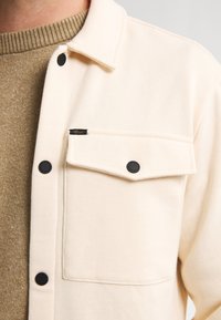 Camisa de botones beige claro con un bolsillo en el pecho, botones negros y una textura suave, llevada sobre un suéter marrón texturizado.