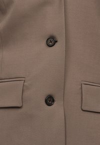 Μπροστινή όψη μπεζ σακακιού με δύο σκούρα κουμπιά και δύο τσέπες με καπάκι, δείχνοντας ομαλό, ελαφρώς υφασμένο ύφασμα.