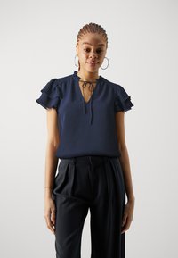 Blusa blu navy con scollatura a V, maniche corte arricciate e dettaglio con fiocco al collo. Indossata con pantaloni neri su misura. Texture liscia.