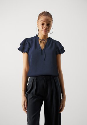 Navy blue blouse with v-neck, ruffled short sleeves, and a tie detail at the neck. Worn with tailored black trousers. Smooth texture.