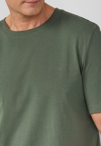 Oliwkowa koszulka z okrągłym dekoltem, wykonana z miękkiej bawełny, z małym haftowanym logo po lewej stronie z przodu. Krótkie rękawy, gładka faktura.