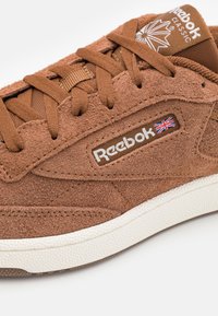 Hnědé semišové tenisky s plochými tkaničkami, logem Reebok a detailemi britské vlajky. Bílé gumové podrážky s texturovaným povrchem, s designem zaoblené špičky.