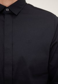 Camisa de cuello navy confeccionada en tejido suave, con un sutil logo A|X bordado en el pecho, con una tapeta de botones y un diseño ajustado.