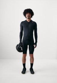 Svart sykkelsett som inkluderer en tettsittende langermet trøye med glidelås og polstrede shorts. Tilbehør: svart hjelm og syklesko.