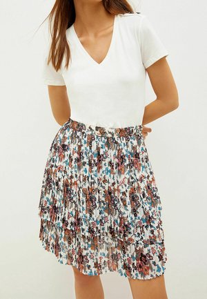 Vrouw draagt een wit T-shirt met V-hals en korte mouwen, ingestopt in een geplooide, gelaagde bloemenrok met bruine, blauwe en roze patronen.