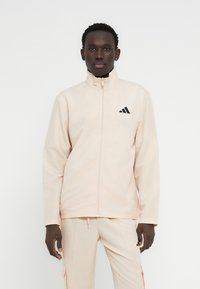 Νεαρός άνδρας που φοράει ανοιχτό μπεζ τζάκετ με φερμουάρ και αντίστοιχο παντελόνι με μαύρα λογότυπα Adidas, στέκεται μπροστά σε απλό λευκό φόντο.