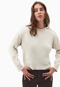 Maglione color crema con collo rotondo e polsini a coste. Presenta bottoni dorati sulle spalle. Design a maglia semplice e texture.