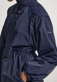 Veste bleu marine en matériau léger, avec un col haut, des poignets élastiques et une taille à cordon de serrage. Logo sur la manche.