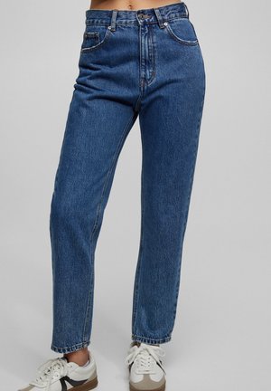 Blauwe recht gesneden jeans met hoge taille, gecombineerd met witte sneakers tegen een effen lichtgrijze achtergrond.