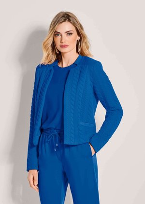 Femme blonde portant une veste bleue texturée sur un haut bleu et un pantalon bleu assorti, debout avec une main dans la poche.