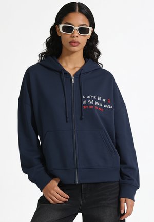 Vrouw met lang donker haar die witte zonnebrillen draagt en een marineblauwe hoodie met rits, met een hart en tekst op de borst.