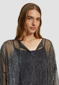 Jeune femme aux longs cheveux châtains clairs portant un haut noir transparent avec des détails scintillants verticaux argentés, sur un fond clair uni.