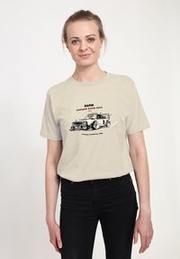 Beige tričko s grafikou závodního auta a textem "RAPID" a "RAPID TRUCK STAR." Černé džíny, jednobarevné pozadí.