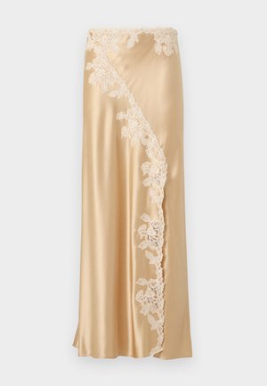 Naine seisab valge seina taustal, kandes kuldse satiini kamisooli ja pikka seelikut pitsist äärisega, beeže sandaale ning rõngaskõrvarõngaid.