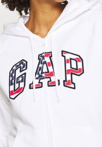Sweat à capuche blanc zippé avec un grand logo "G.A.P" en rouge, blanc et bleu étoilé, doté de cordons de serrage et d'un tissu doux et texturé.