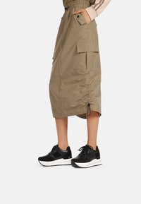 Gonna midi khaki con vita elastica e coulisse, dotata di tasche cargo laterali. Abbinata a sneakers nere con suole bianche.