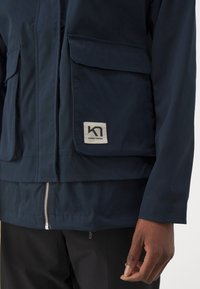 Navy blue jacket featuring a smooth texture, multiple front pockets, a silver zipper, and a rectangular logo patch on the front.