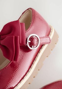 Scarpe in vernice rossa con fiocco a righe e dettaglio della fibbia argentata. Presentano una suola beige antiscivolo e una finitura liscia.