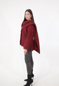 Maglione oversize bordeaux con trama, colletto drappeggiato e maniche ampie, abbinato a pantaloni aderenti grigi e stivali neri con tacco alto.