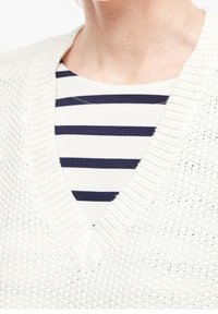 Pull blanc en maille avec un col en V, présentant une surface texturée. En dessous, un haut rayé bleu marine et blanc.
