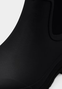 Zwarte rubberen laars met een gestructureerd mat oppervlak, gebogen vorm en een brede opening voor gemakkelijk dragen. Voorzien van een zachte zool en versterkte randen.