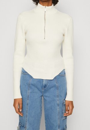 Cream ribbed long-sleeve top with a high collar and half-zip front. Features a cropped, scalloped hem for structured shape.