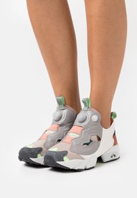 Baskets de sport grises avec dessus en maille, accents multicolores en pêche, vert et marron, dotées d'une fermeture à pompe distinctive et d'une semelle texturée.