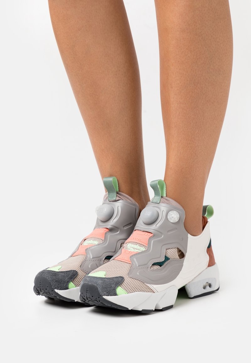 Baskets de sport grises avec dessus en maille, accents multicolores en pêche, vert et marron, dotées d'une fermeture à pompe distinctive et d'une semelle texturée.