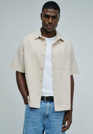 Homme portant une chemise beige à manches courtes boutonnée sur un t-shirt blanc, un jean bleu et une ceinture noire, debout avec une main dans la poche.