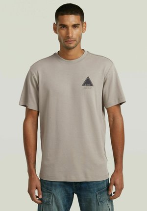 Man met kort haar die een beige G-Star T-shirt draagt met een klein zwart driehoekig logo op de borst en blauwe cargobroek, staand tegen een effen achtergrond.
