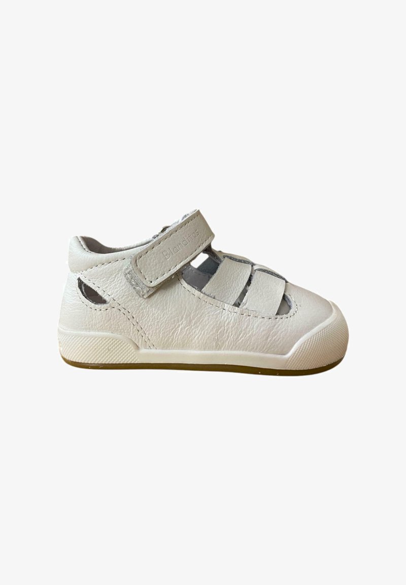 Zapato de cuero blanco con una correa de velcro, lados abiertos, punta redondeada y suela texturizada. Presenta un sutil logotipo en la correa.