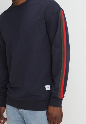 Homme portant un sweat-shirt bleu marine foncé avec des rayures rouges et vertes sur la manche et un jean bleu clair, debout devant un fond uni.