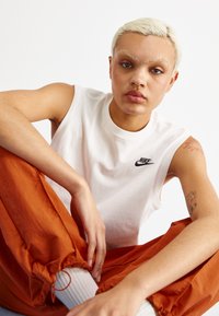 Camisa sin mangas blanca de Nike, pantalones naranjas con puños elásticos, tejido texturizado y tatuajes visibles en los brazos.