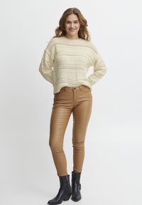 Maglione lavorato a maglia crema con motivo traforato, abbinato a pantaloni in ecopelle marroni aderenti e stivaletti neri alla caviglia. Design semplice, stile casual.