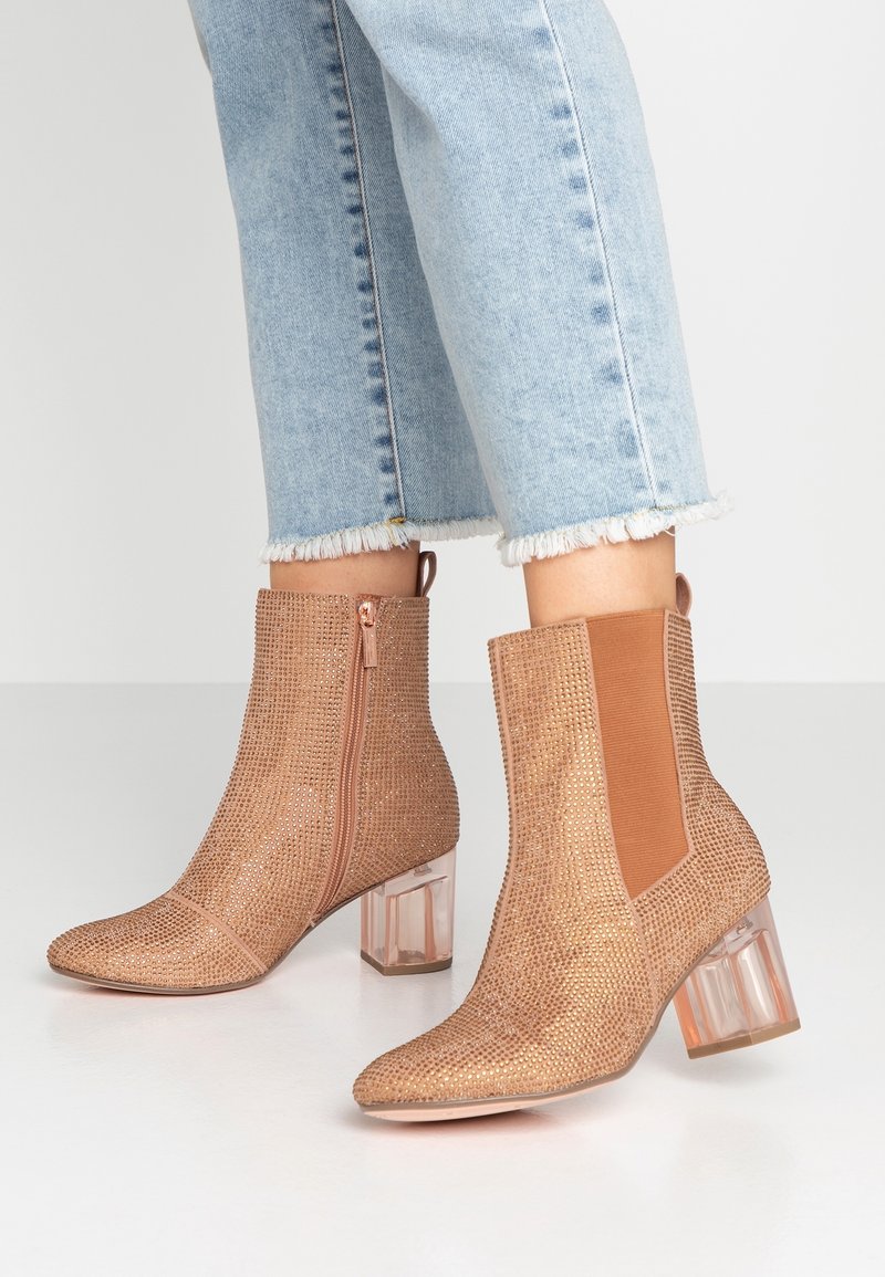 Botas de tobillo en oro rosa con una superficie de malla texturada, tacones de bloque transparentes, cremallera lateral y paneles elásticos; combinadas con unos pantalones vaqueros recortados de color azul claro.