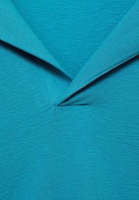 Turkizna, teksturirana tkanina s ovratnikom i umetnutim, kutnim dizajnom sprijeda, s blagim valovitim uzorcima.