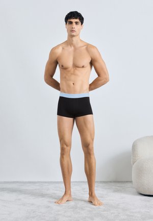 Černé boxerky s lehce modrým pasem, které mají těsný střih. Model stojí bos na světle šedém koberci před minimalistickým pozadím.