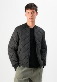Giovane uomo con capelli corti e scuri, che indossa una giacca bomber trapuntata nera, una camicia nera e pantaloni verde oliva, in piedi contro uno sfondo bianco.