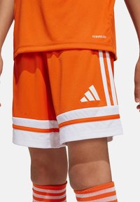 Orangen Sportshorts mit weißen Streifen und Akzenten. Sie besitzen eine einfarbige Optik, eine glatte Textur und ein Adidas-Logo an der unteren Seite.