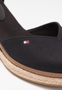 Chaussure en tissu noir avec semelle tressée beige et petit logo rectangulaire rouge, blanc et bleu sur le côté supérieur près de la couture.