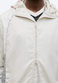 Man i en ofodrad beige jacka över svarta och vita lager av toppar, med en silverkedja synlig runt halsen.