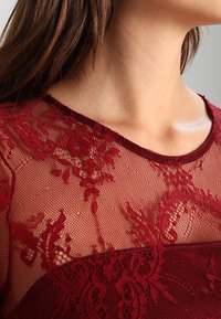 Burgundy lace top with floral pattern and textured mesh, featuring a round neckline and short sleeves, highlighting intricate detailing.